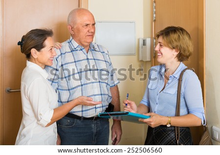Mature couple answer questions of smiling woman with papers at door in home. Focus on man