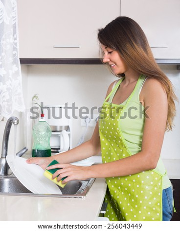 Young housewife in apron washing plates with sponge in domestic kitchen
