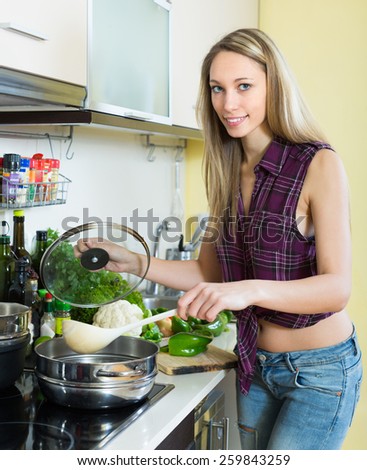 Smiling blonde woman with ladle testing soup from pan in domestic kitchen