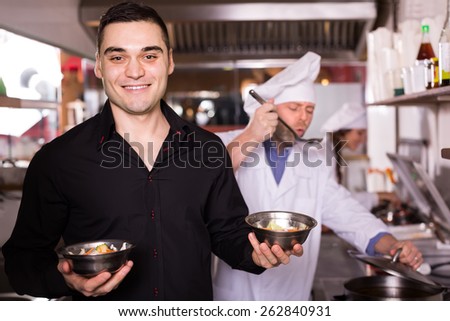 Smiling handsome male waiter taking away prepared food at bistro kitchen