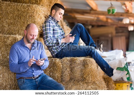 Two farmers resting on a stack of straw during their work day on a farm playing on smartphones