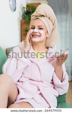 Positive woman in bathrobe putting cosmetic treatment on face in house