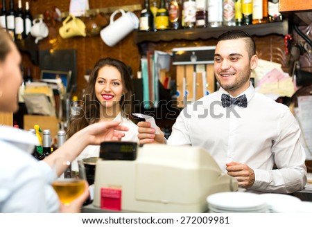 Joyful staff working in a bar: female bartender is smiling, male barista is handing over a check to a waiter