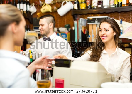 People working in a bar: waitress is taking a check from a cashier while handsome bartender is mixing drinks in background