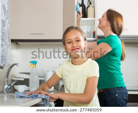 Smiling girl helping mother dusting furniture indoor. Focus on girl