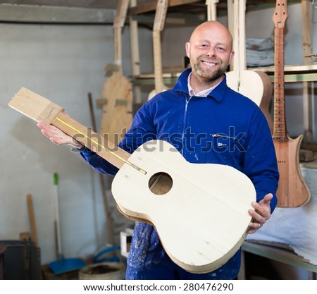 Portrait of happy professional guitar maker holding unfinished guitar indoors