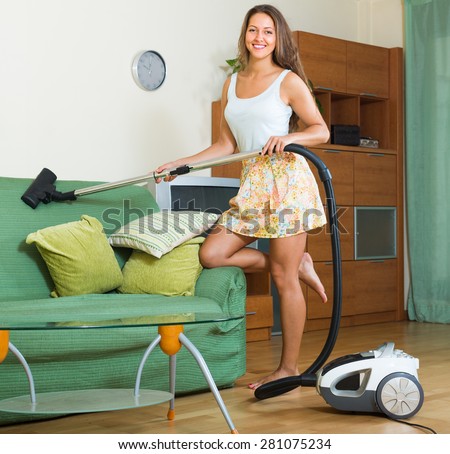 Smiling young woman in skirt cleaning living room with vacuum cleaner