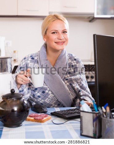 Female freelancer in bathrobe with PC, tea and sandwich at kitchen table