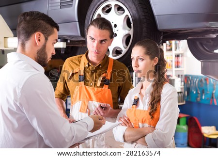 Unhappy client talking to a repair crew in a car shop. Car is hanging on a lift in background
