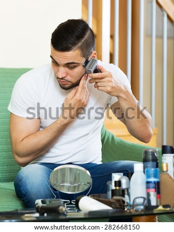 Beautiful adult man shaving electric shaver at home sitting at a sofa in the living room