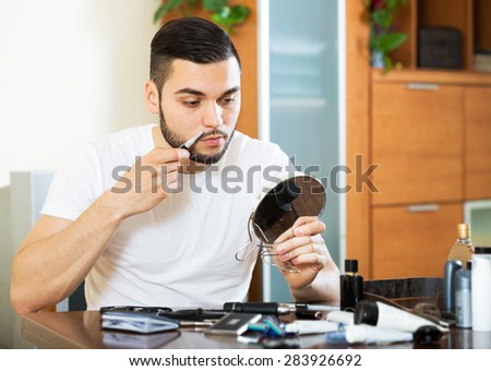 Handsome man shaving electric shaver at home sitting at a table in the living room