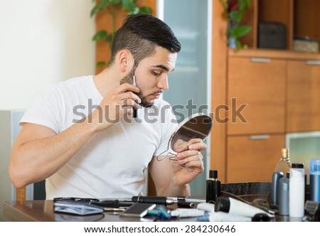 young european guy looking at mirror and shaving beard with trimmer