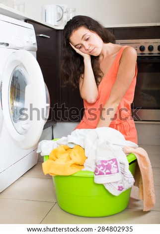 Sad young housewife with clothes near washing machine at home