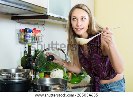 Smiling young blonde woman with ladle testing soup from pan in domestic kitchen