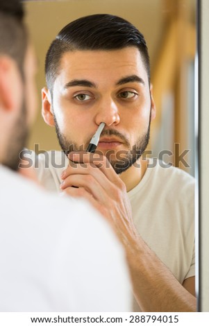 Young man using trimmer for removing hair in his nose in front of a mirror