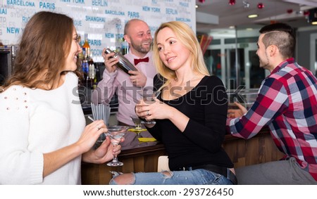Happy girlfriends are having a conversation at a bar while barman is mixing a cocktail in a shacker for a man on the background