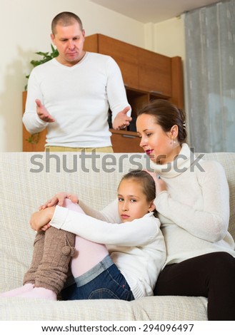 Sad mother comforting litlle daughter in livingroom and angry father