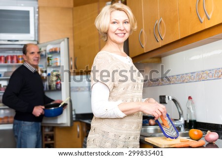 Portrait of senior couple cooking in the kitchen. Beautiful elderly woman is grating carrots while her husband is taking a pan from an opened refridgerator