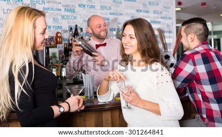 Two women and a man at the bar. Women are talking and a man is waiting for his drink while the bartender is mixing it in a shacker