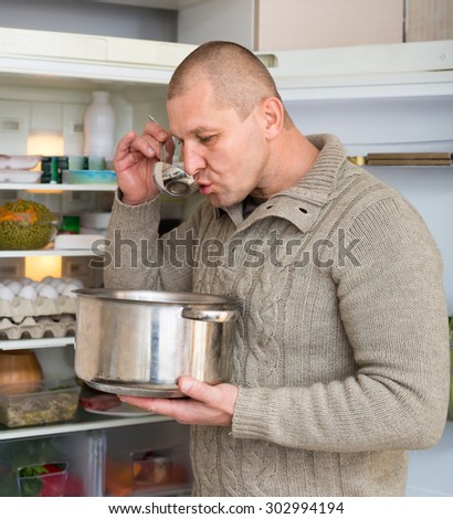 Hungry man eating soup from pan near fridge at home