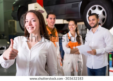 Satisfied client showing thumbs up at a repair shop. Lifted up car and repair crew standing and smiling in the background