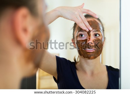 Young smiling woman applying mask on her face in front of mirror