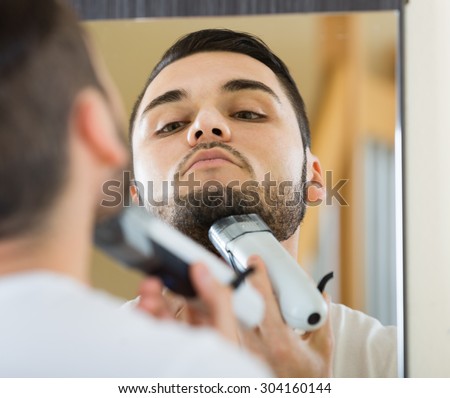 russian man looking at mirror and shaving beard with trimmer