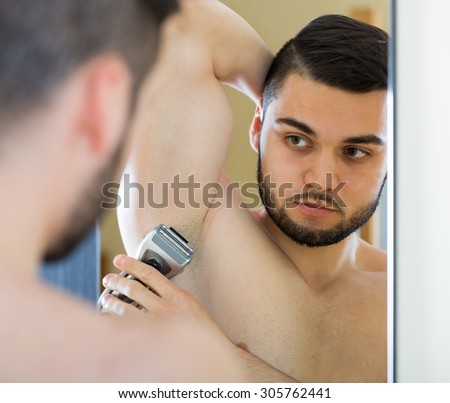 Spanish man looking at mirror and shaving armpit with trimmer