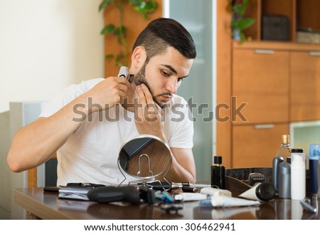 positive american man looking at mirror and shaving beard with trimmer