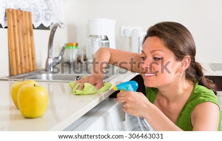 Cheerful girl in green cleaning furniture in kitchen at home
