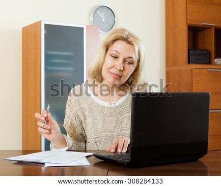 Serious mature woman staring financial documents with laptop  at table in office interior
