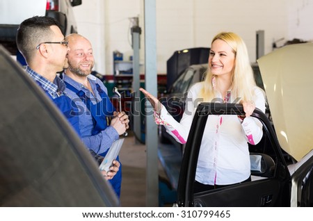 Satisfied female customer is taking her car from a repair shop after maintenance done by two handsome smiling mechanics