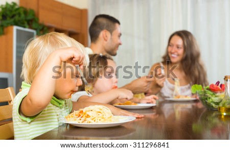 Happy young parents with children having lunch with spaghetti at home together. Focus on girl