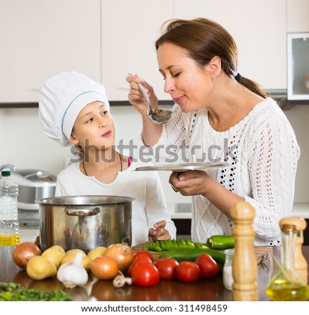 Portrait of cheerful daughter and mom with vegetables and casserole in kitchen at home. Focus on girl