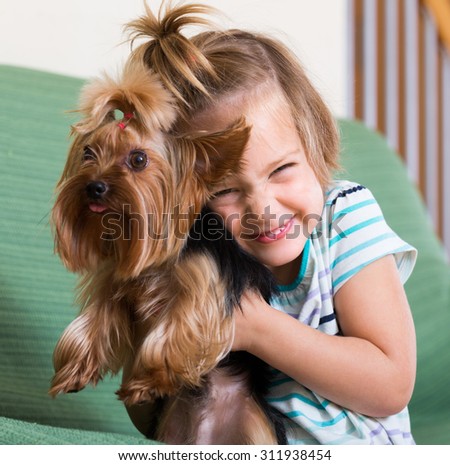 Happy cute blonde little girl playing with Yorkshire Terrier on sofa indoor