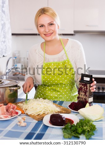 Cheerful blonde woman chopping cabbage for Russian soup at kitchen table