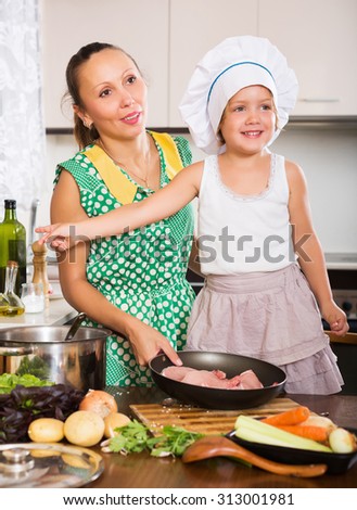 Happy woman with baby in cook hat cooking with meat at kitchen