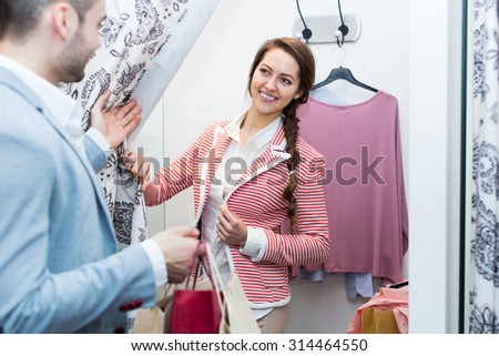 Positive young couple with new apparel at fitting-room in clothing store