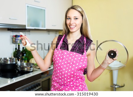 Beautiful smiling woman cooking in the kitchen. She is holding a cap of a pan and a spoon in her hands