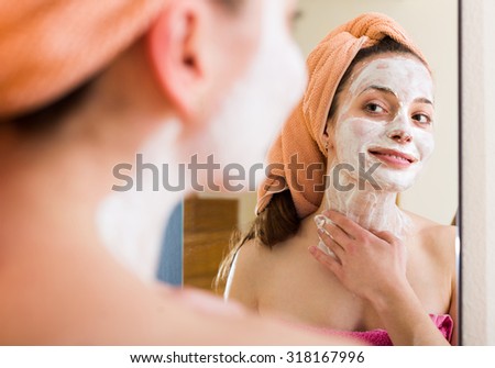 Young woman in bathrobe applying face pack in front of the mirror indoors