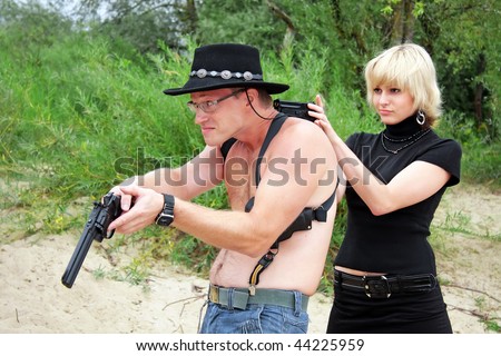 woman pointing gun at surrendering man with revolver wearing cowboy hat, sand and trees on background