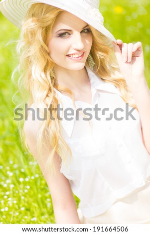 Portrait of a beautiful young blonde woman with wavy hair in nature. Girl in white hat sitting on the grass in the park