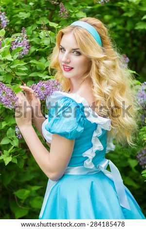 Portrait of a beautiful young blonde woman with long hair dressed as Alice in Wonderland. Girl on the nature near the lilac bushes. Soft focus