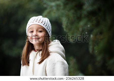Adorable little girl posing near the branch of tree and smiling in to a camera. Wearing winter coat and hat. Lovely young girl in the winter outdoors.