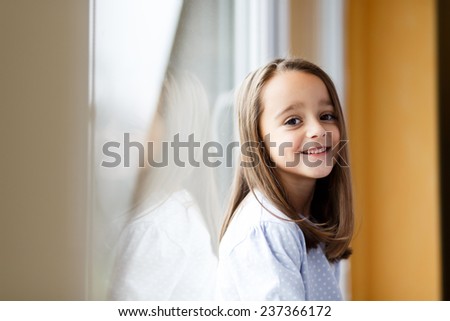 Beautiful little girl smiling and watching out the window. A child looks out the window. Young girl looking from window. Portrait of cheerful kid sits at windowsill.