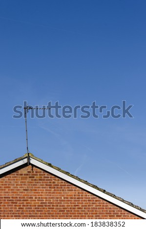 TV antenna / aerial at apex of sloping tiled roof on top of red brick wall against mainly blue sky with hazy clouds.