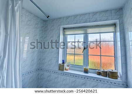 Wide angle bathroom interior, inc edge of shower curtain and curtain rail, tiled walls, frosted glass window with distorted exterior, and various items on the windowsill [landscape format].