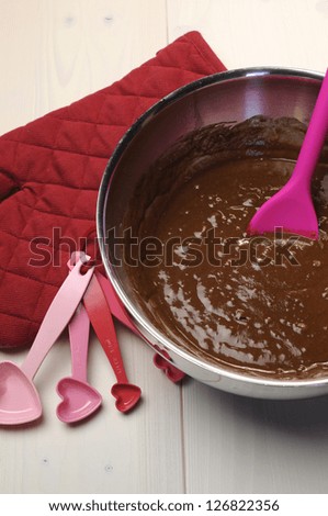 Preparation of cake baking with chocolate batter in steel bowl, red glove and heart shape measuring spoons on vintage shabby chic natural white wood table.