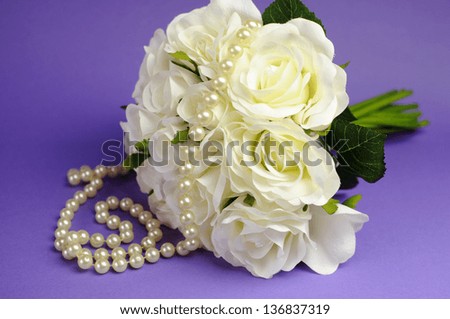 Wedding bouquet of white roses with string of pearls necklace, against purple lilac background.