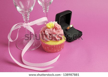 Wedding theme bridal pair of champagne flute glasses with pink cupcake and wedding ring in black jewelry box against a pink background.